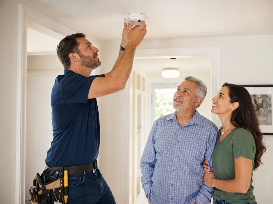 A vetted provider installing a smoke detector while a homeowner couple looks on